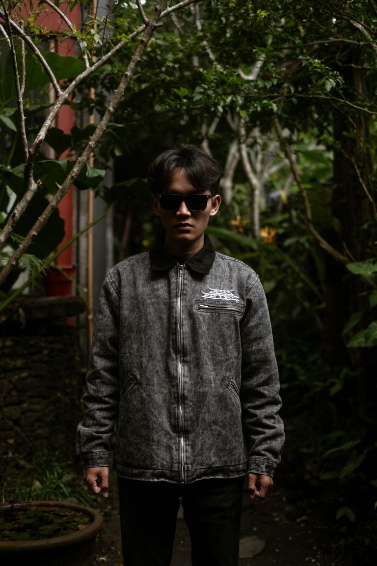 A young man in stylish sunglasses and denim jacket standing in an urban garden area.